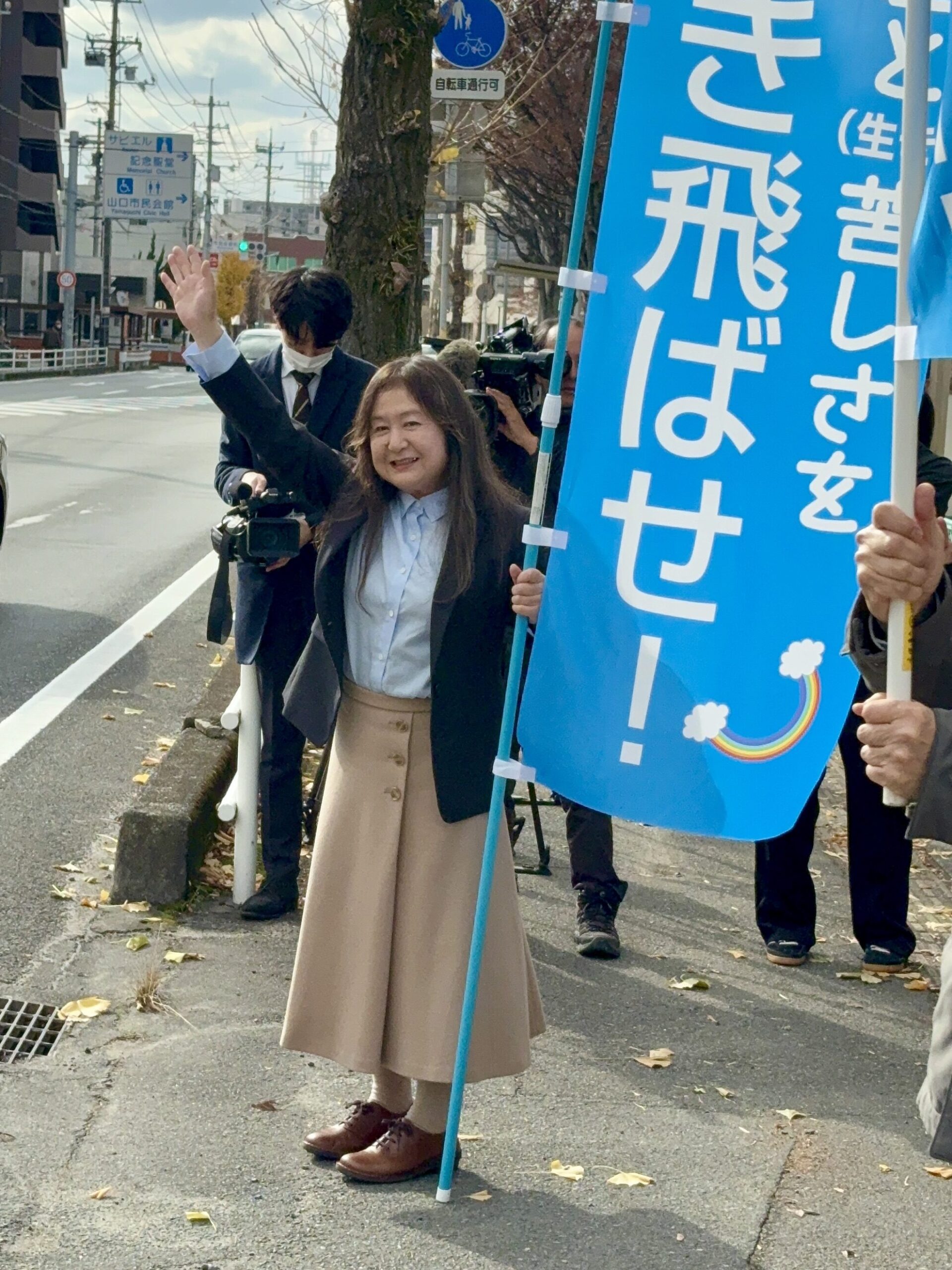事務所前で息(生き)苦しさを吹き飛ばせ!と書かれた旗を持って手を振る大久保さん